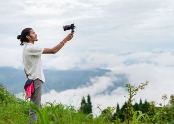 A young man films a lush, misty mountain landscape on an overcast day, capturing nature's beauty.