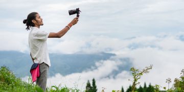 A young man films a lush, misty mountain landscape on an overcast day, capturing nature's beauty.