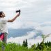 A young man films a lush, misty mountain landscape on an overcast day, capturing nature's beauty.