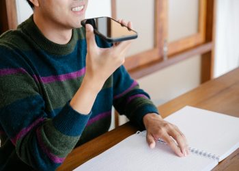 Adult man using smartphone for voice commands, smiling at home desk.