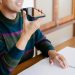 Adult man using smartphone for voice commands, smiling at home desk.