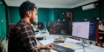 Software developer coding on dual monitors in a well-lit modern office, focused and engaged.