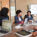 Four diverse women engaged in a business meeting with laptops and presentations in an office.