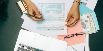 Hands writing on tax documents with laptop, glasses, and currency on desk.