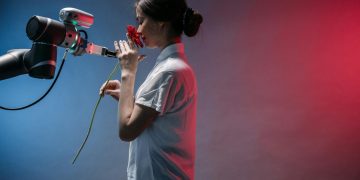 A woman in a lab coat interacts with a robot arm holding a red flower, symbolizing technology and nature.