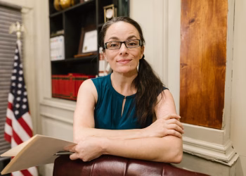 Woman in a blue top with glasses smiling in an office with an American flag.