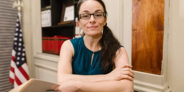 Woman in a blue top with glasses smiling in an office with an American flag.