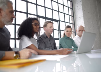 From below of focused multiracial colleagues of different ages in casual wear surfing netbook while making project together in light workplace