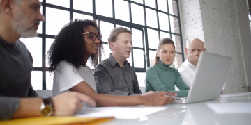 From below of focused multiracial colleagues of different ages in casual wear surfing netbook while making project together in light workplace