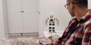 A man examines a robotic toy placed on a table in a contemporary bedroom setting.
