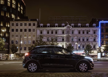 A modern car parked on a city street at night, surrounded by illuminated buildings in an urban downtown area.