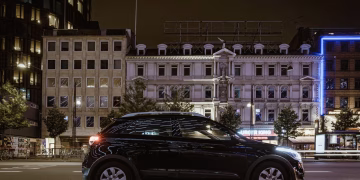 A modern car parked on a city street at night, surrounded by illuminated buildings in an urban downtown area.