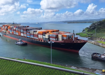 Large container ship maneuvered by tugboats through the Panama Canal under a clear sky.