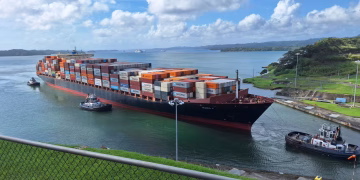 Large container ship maneuvered by tugboats through the Panama Canal under a clear sky.
