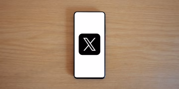 Minimalist image of smartphone with X logo on wooden surface, showcasing digital branding.