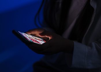 A teenager browses a smartphone in a dimly lit room, highlig