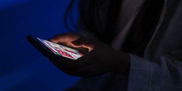 A teenager browses a smartphone in a dimly lit room, highlig