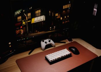 Close-up of a gaming desk setup featuring a keyboard, controller, and monitor at night.