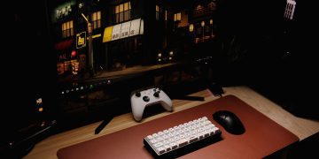 Close-up of a gaming desk setup featuring a keyboard, controller, and monitor at night.