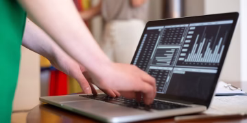 Hands typing on a laptop displaying data charts in an indoor setting.