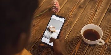 An individual using a dating app on a smartphone with a cup of tea nearby on a wooden table.