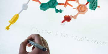 Anonymous chemist writing with marker on illuminated magnetic board formula of Hydronium and Acetate production after studying plastic model of molecule in laboratory