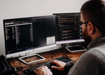 Software developer typing code on dual monitors at a wooden desk.