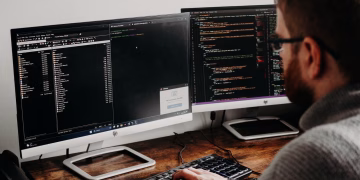 Software developer typing code on dual monitors at a wooden desk.