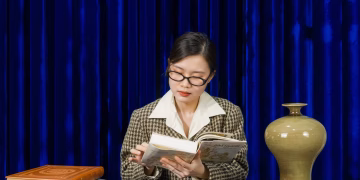 A young woman reading a book at a table with a classic decor vase; set against a dark blue curtain.