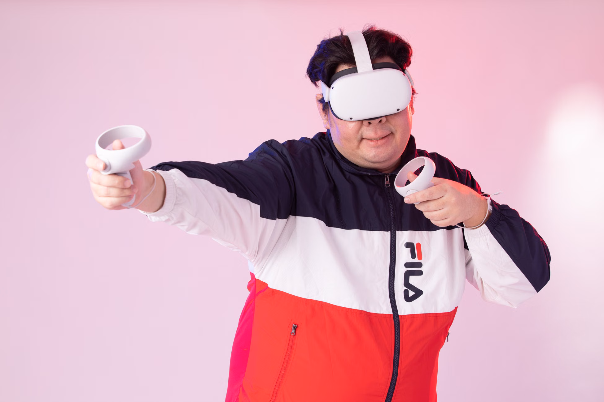 Adult man wearing VR goggles and jacket, playing with controllers against a pink studio background.