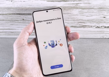 A close-up of a hand holding a smartphone displaying the One UI 6.1 welcome screen indoors.