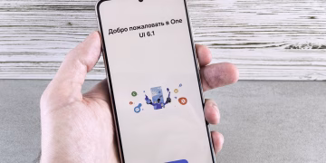 A close-up of a hand holding a smartphone displaying the One UI 6.1 welcome screen indoors.