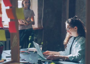 Two women working together in a sunlit office space with a laptop and notes.