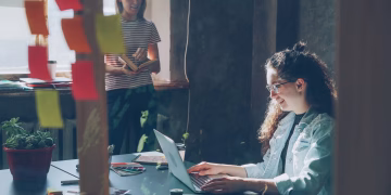 Two women working together in a sunlit office space with a laptop and notes.