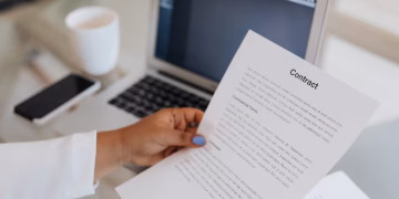 Close-up of a person holding a contract document next to a laptop during a work session.