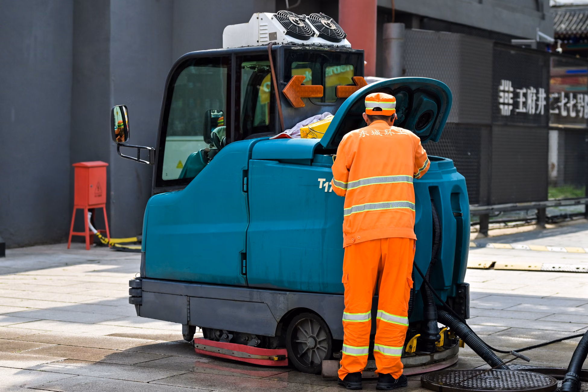 A street cleaner in uniform operates a cleaning machine on an urban sidewalk.