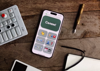Smartphone displaying an app next to an OP-1 synthesizer, pen, and notebook on a wooden surface.