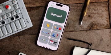 Smartphone displaying an app next to an OP-1 synthesizer, pen, and notebook on a wooden surface.