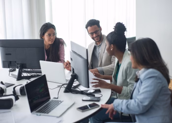 A diverse team engages in discussion around computers in a modern office setting.