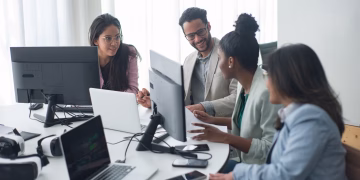 A diverse team engages in discussion around computers in a modern office setting.