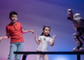 Two children joyfully dance with a robot in a colorful indoor setting.