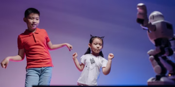 Two children joyfully dance with a robot in a colorful indoor setting.