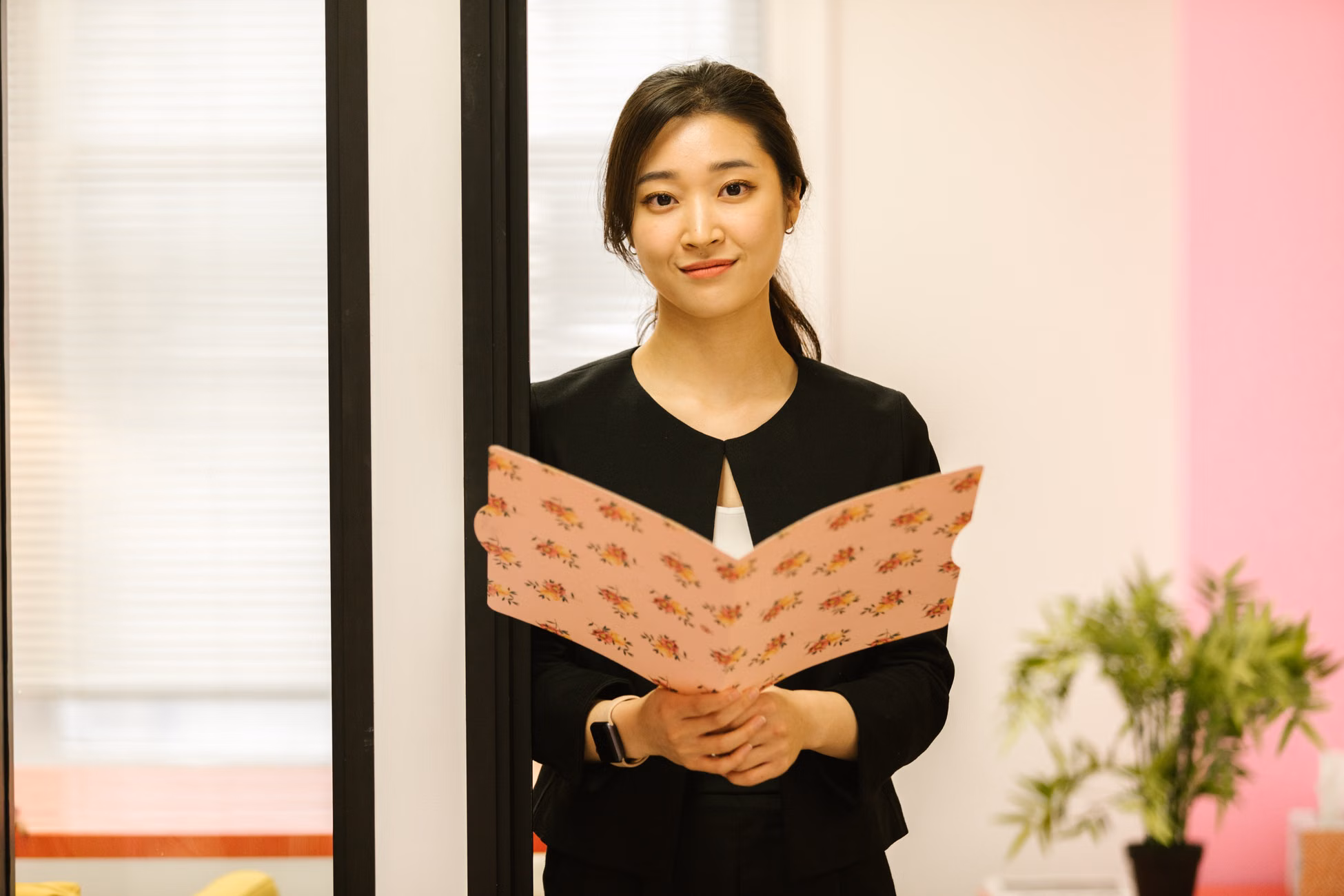 Confident woman in office attire holding a floral folder, standing in a modern workplace.