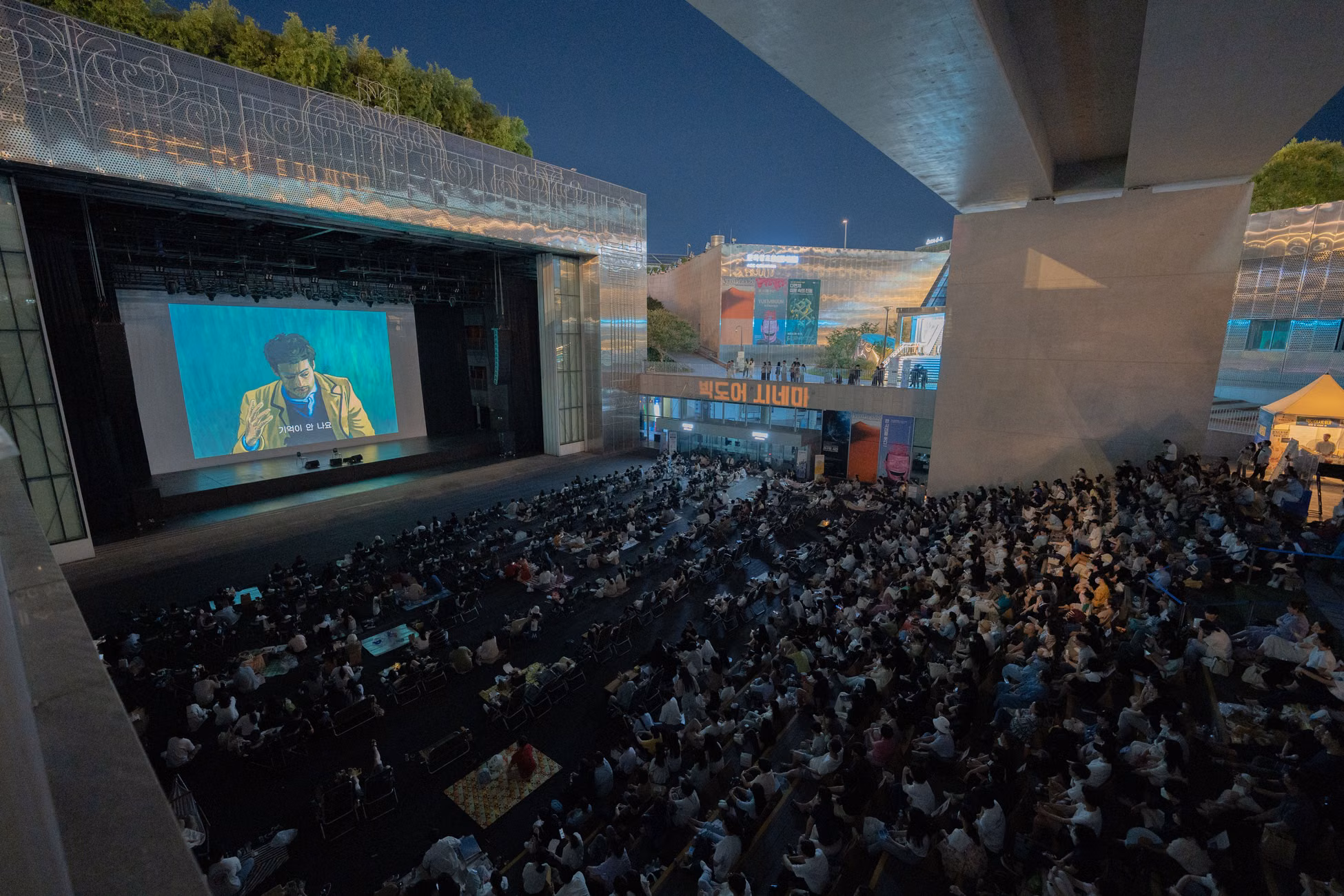 A large audience gathers outdoors at night to watch a movie screening under a bridge.