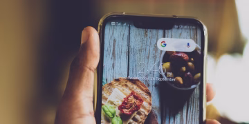 Close-up of a hand holding a smartphone displaying a food image on its screen, indoors.