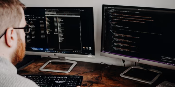 Software developer working on code with dual monitors in a home office setting.