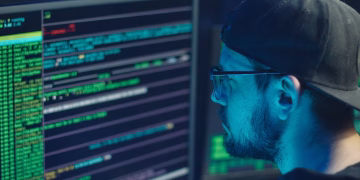 A cybersecurity expert inspecting lines of code on multiple monitors in a dimly lit office.