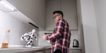 Asian man smiling while playing with a robot in a modern kitchen setting.
