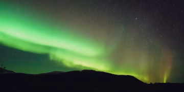From below picturesque view of colorful shiny polar lights illuminating sky over mount silhouette at dusk