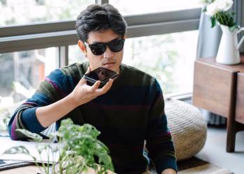 A man wearing sunglasses uses a smartphone's voice assistant in a cozy indoor setting.